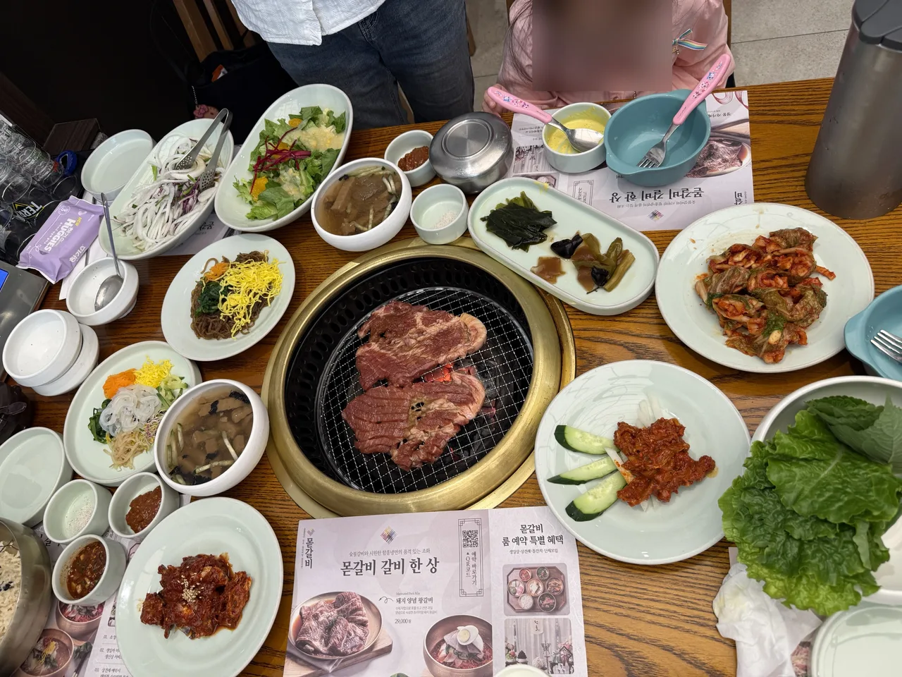 숯불 불판을 가운데 두고 잡채와 샐러드, 김치, 냉면 육수가 차려진 몯갈비 상차림, 아이 얼굴은 가림